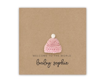 May include: A brown card with the text "Welcome to the world baby Sophie" and a pink crocheted baby hat with a white pom-pom on top.