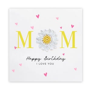 May include: White birthday card with the word "MOM" in yellow, a crocheted daisy, and the text "Happy Birthday I LOVE YOU". Pink hearts are scattered around the card.