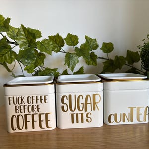 Puede incluir: Tres botes de cocina de cerámica blanca con ribete y tapas doradas. Los botes tienen letras doradas que dicen "FUCK OFFEE BEFORE COFFEE", "SUGAR TITS" y "CUNTEA". Hay follaje verde en el fondo.