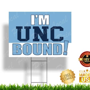 May include: A blue and white yard sign with the text "I'M UNC BOUND!" in navy blue letters. The sign is on a metal stake and has a blue background with white lettering. The sign is surrounded by green grass.