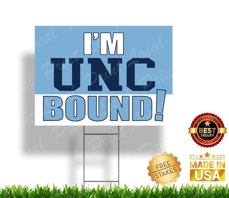 May include: A blue and white yard sign with the text "I'M UNC BOUND!" in navy blue letters. The sign is on a metal stake and has a blue background with white lettering. The sign is surrounded by green grass.