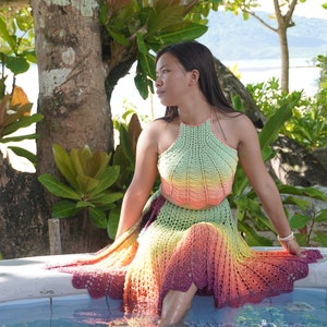 May include: A woman wearing a crocheted halter top and skirt set in a rainbow ombre pattern. The set is made of a soft, lightweight yarn and features a delicate crochet stitch. The woman is sitting on the edge of a pool, with lush greenery in the background.