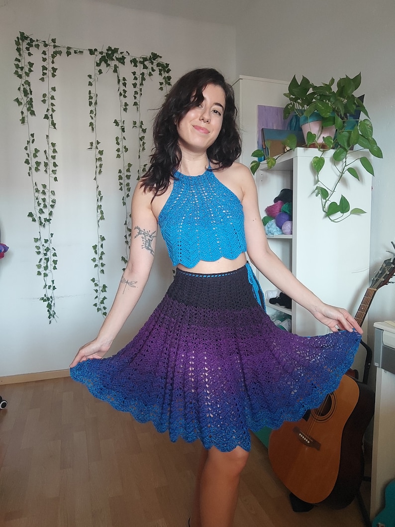 May include: A woman wearing a blue crochet halter top and a purple and blue ombre crochet skirt. The skirt is a-line and has a scalloped hem.