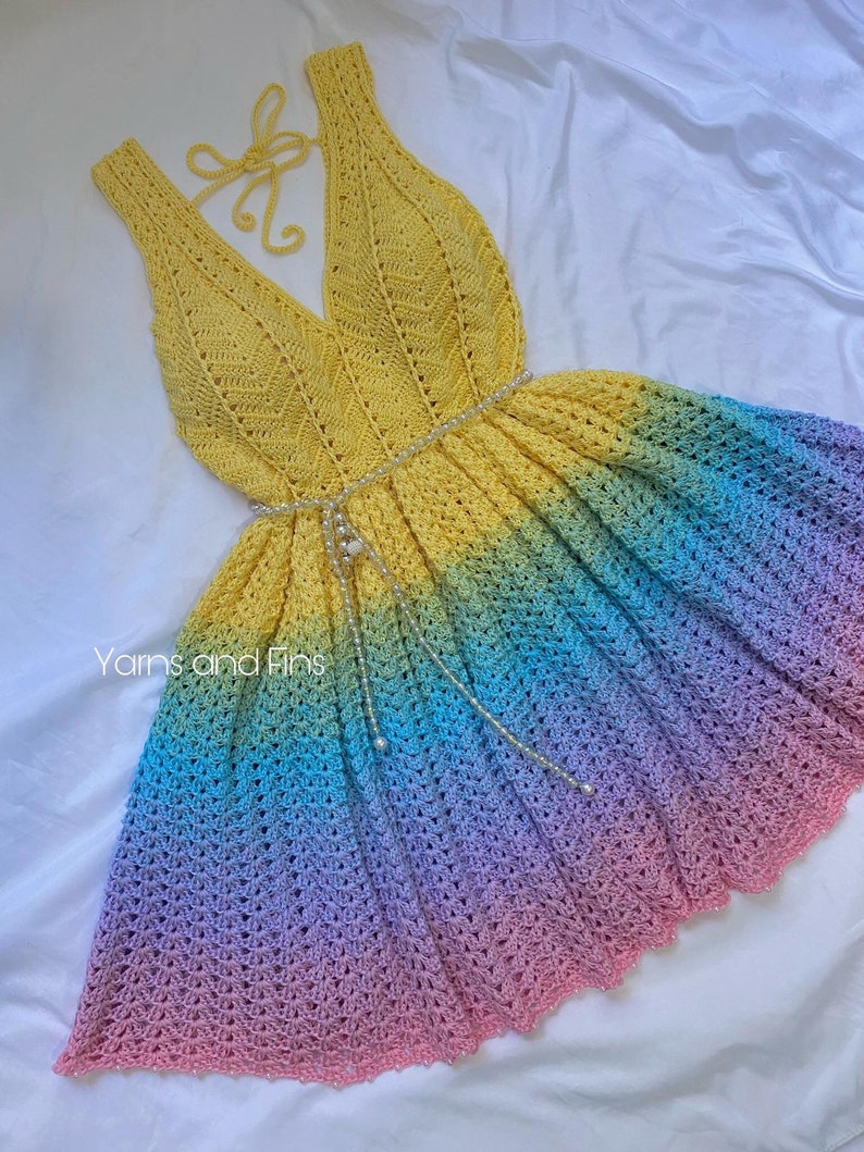 May include: A yellow crochet dress with a rainbow ombre skirt. The dress has a V-neckline and a halter strap. The dress is decorated with a pearl-like beaded belt.