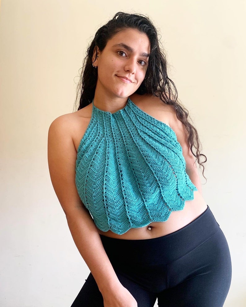 Op de afbeelding: Een turquoise haltertop met geschulpte rand. De top is gemaakt van gehaakt materiaal. Het model draagt een zwarte legging. Een uniek en stijlvol kledingstuk.