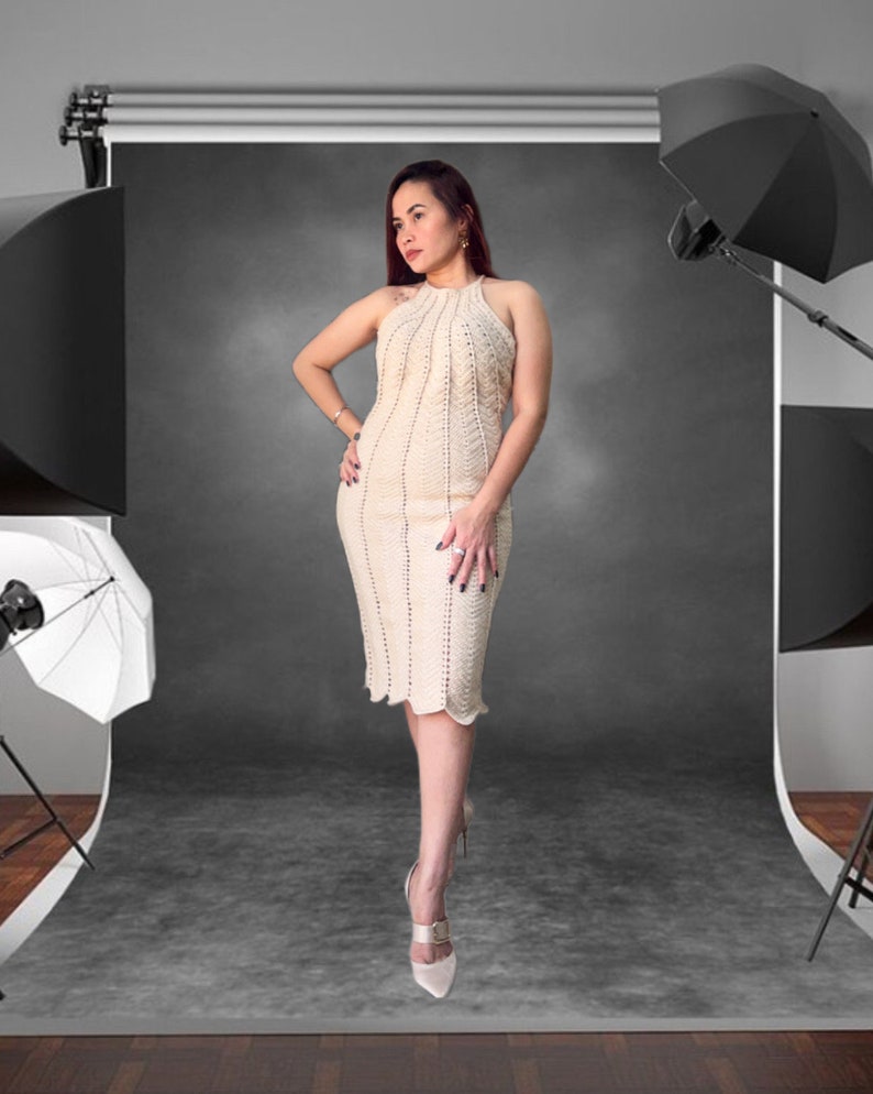 May include: A woman wearing a white crocheted dress with a halter neckline and a beaded design. She is standing in a studio setting with a black backdrop and lighting equipment.