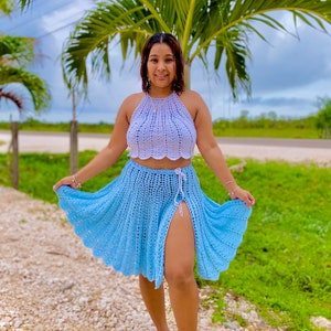 May include: A woman wearing a light blue crochet skirt with a side slit and a white crochet halter top. She is standing on a path with a palm tree in the background.