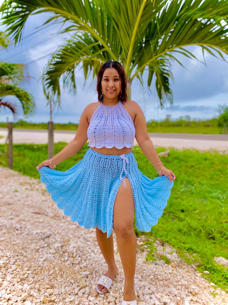 May include: A woman wearing a light blue crochet skirt with a side slit and a white crochet halter top. She is standing on a path with a palm tree in the background.