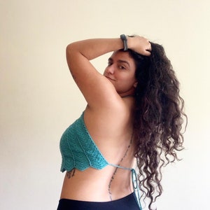 Op de afbeelding: Een turquoise gehaakte haltertop met een geschulpte rand. De top wordt op de rug vastgebonden met een turquoise touwtje. Het model draagt een zwarte broek. De achtergrond is effen cr&egrave;mekleurig.