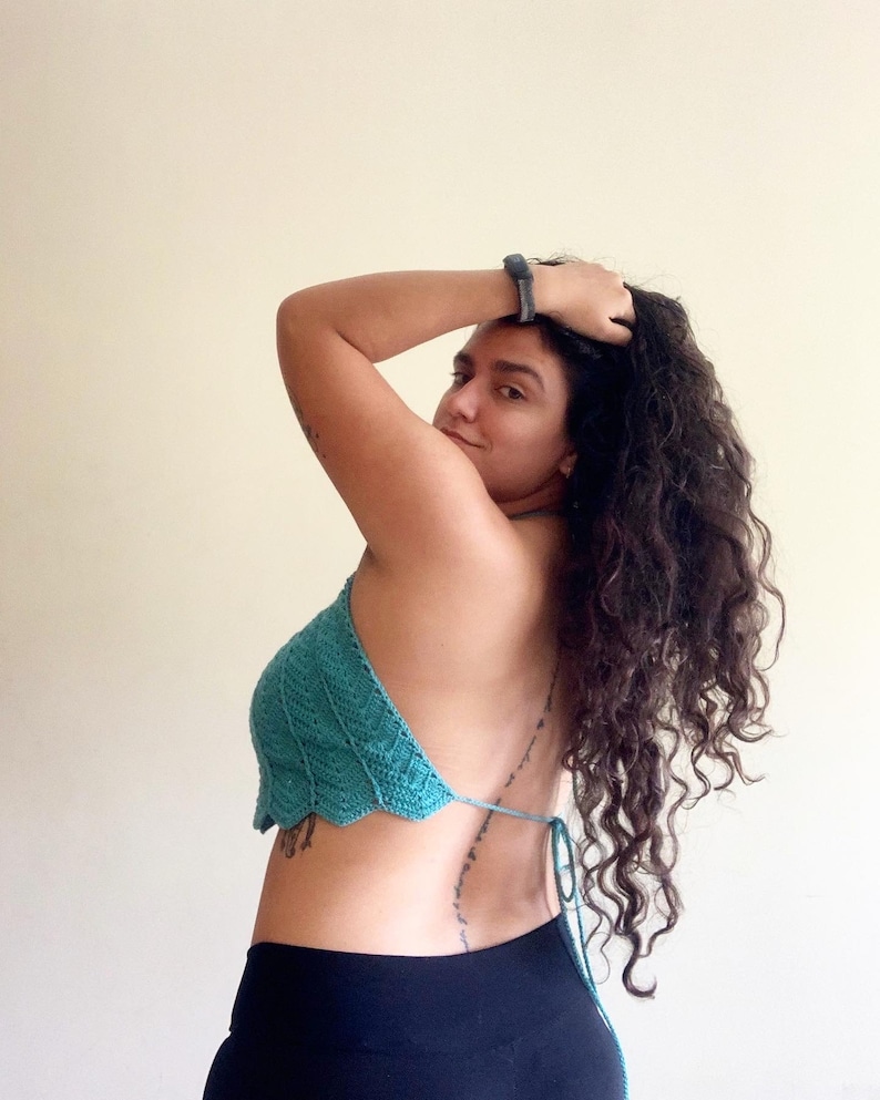 Op de afbeelding: Een turquoise gehaakte haltertop met een geschulpte rand. De top wordt op de rug vastgebonden met een turquoise touwtje. Het model draagt een zwarte broek. De achtergrond is effen cr&egrave;mekleurig.
