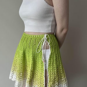 May include: A green crochet skirt with a white crochet trim. The skirt is tied at the waist with a white string and has a slit up the side.