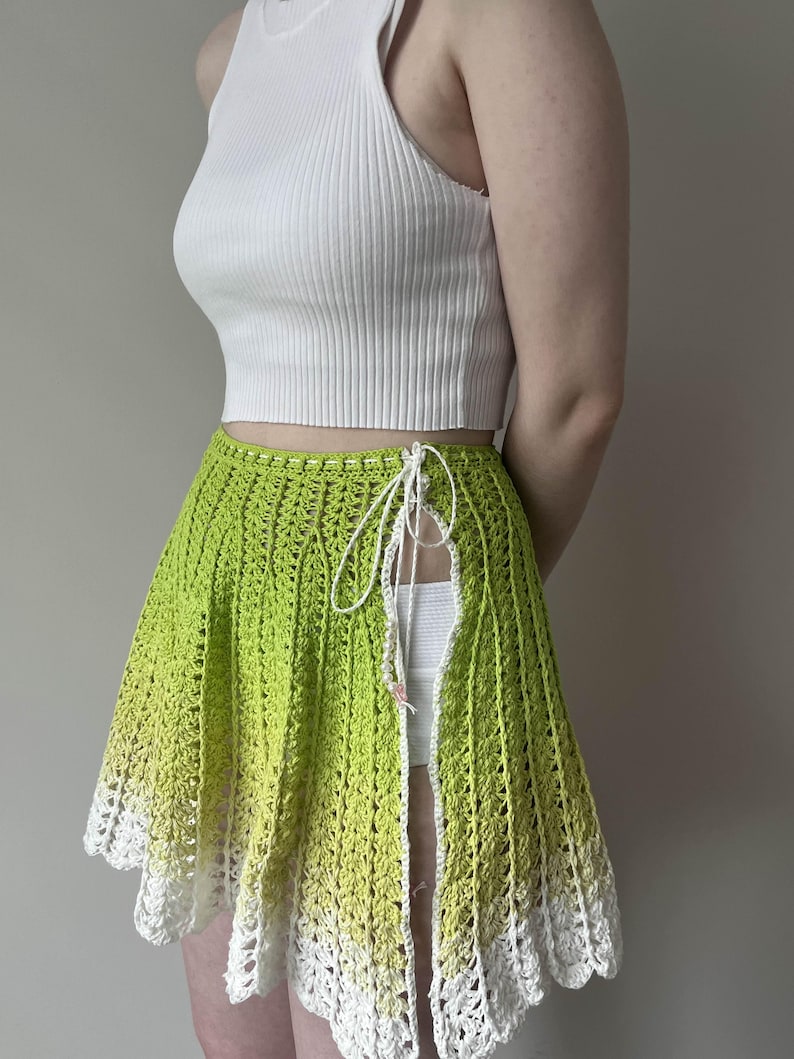 May include: A green crochet skirt with a white crochet trim. The skirt is tied at the waist with a white string and has a slit up the side.