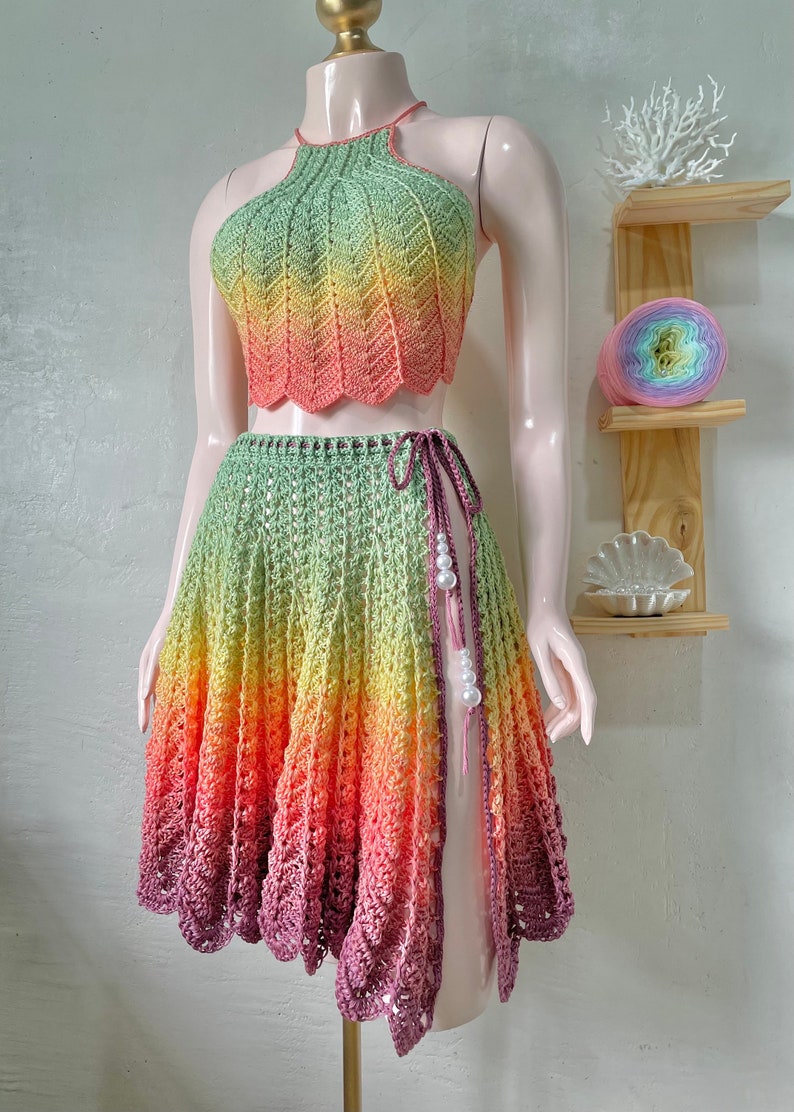 May include: A crochet skirt and halter top set in a rainbow ombre pattern. The skirt has a side slit and is tied at the waist with a matching cord. The top has a halter neckline and is crocheted in a chevron pattern.