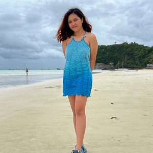 May include: A woman wearing a blue crochet halter dress and sandals stands on a sandy beach. The sky is cloudy and the sea is in the background.