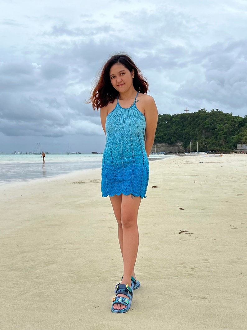 May include: A woman wearing a blue crochet halter dress and sandals stands on a sandy beach. The sky is cloudy and the sea is in the background.