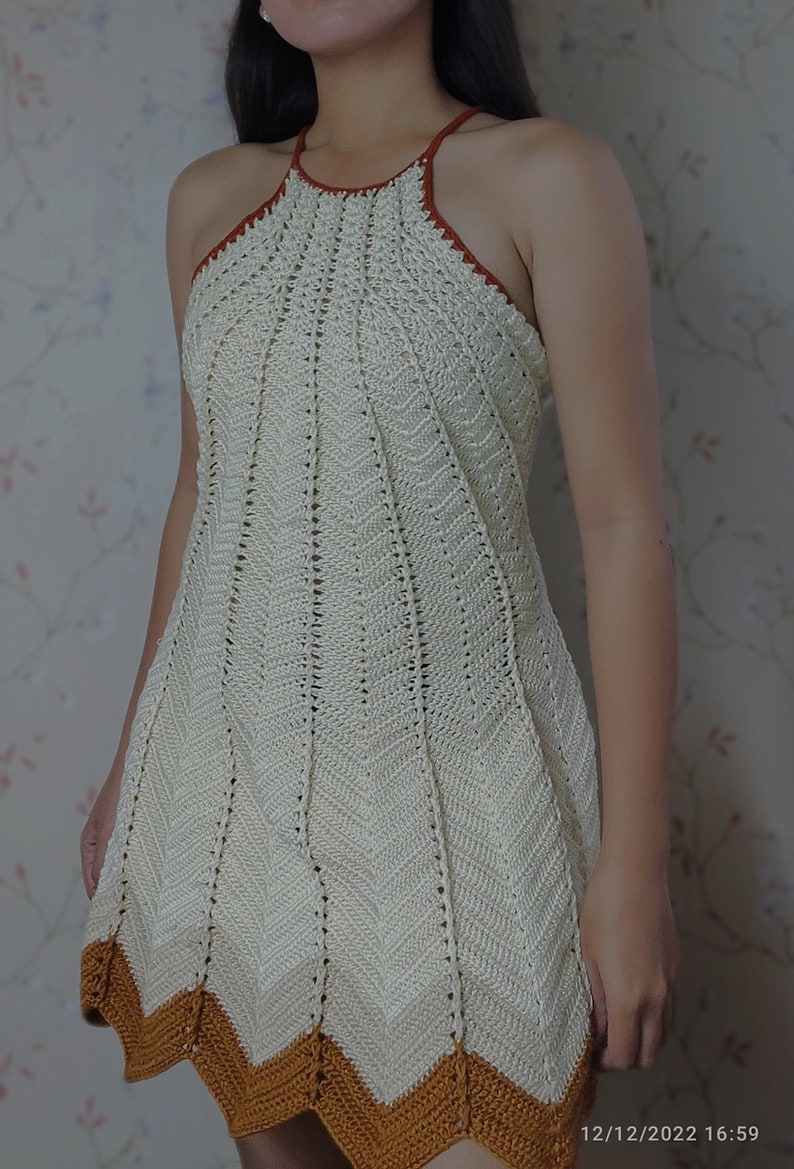May include: A white crochet halter dress with a brown crocheted trim. The dress has a fitted bodice and a flared skirt.