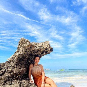 May include: A woman wearing a rust-coloured crochet dress sits on a large rock formation on a beach. The sky is blue with white clouds and the ocean is turquoise.