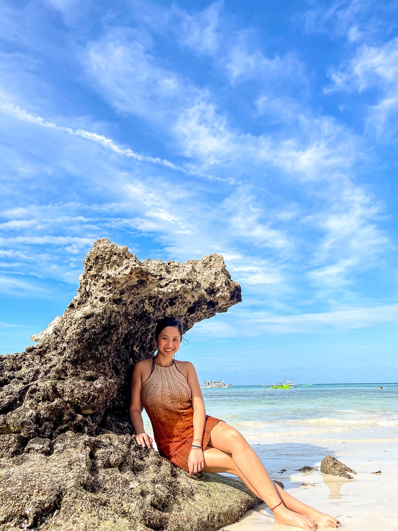 May include: A woman wearing a rust-coloured crochet dress sits on a large rock formation on a beach. The sky is blue with white clouds and the ocean is turquoise.