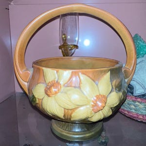 May include: A ceramic basket with a large handle, featuring a floral design with yellow and orange flowers. The basket has a brown base and rim, and a clear glass object is visible behind it.
