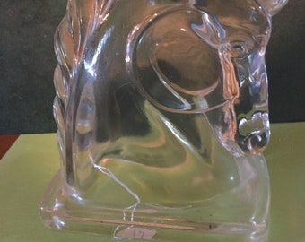 May include: A clear glass horse head bookend. The horse's head is facing to the left and the bookend has a rectangular base.