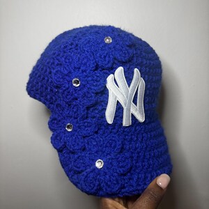 Op de afbeelding: Een koningsblauwe gehaakte baseballpet met een wit "NY" logo en drie heldere strass-steentjes. De pet heeft een gestructureerd, bloemachtig patroon en een gebogen klep. De pet wordt door een hand vastgehouden.