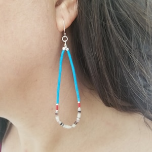 May include: Teardrop-shaped earrings with turquoise blue beads and silver hooks. The earrings feature a mix of white, black, and red beads at the bottom. These handmade earrings are a unique accessory.