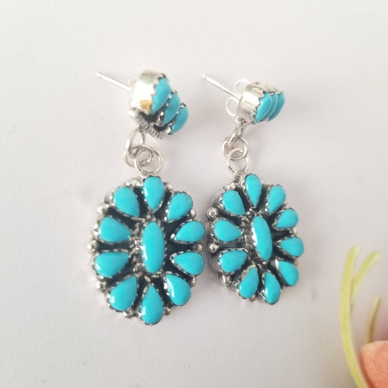 May include: A pair of silver dangle earrings with turquoise stones. The earrings feature a cluster of turquoise stones in the shape of a flower.