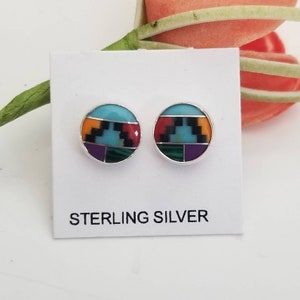 May include: A pair of sterling silver earrings with turquoise, red, yellow, black, green, and purple inlay. The earrings are round and have a geometric design.