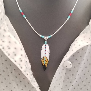 May include: A silver chain necklace with turquoise and red beads. The necklace has a pendant in the shape of a white feather with black, yellow, and turquoise accents.