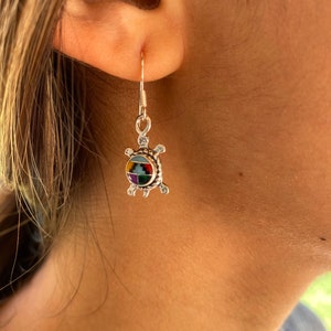 May include: A silver turtle-shaped earring with a colorful mosaic design. The earring is suspended from a silver hook and is worn on a person's ear. The mosaic features a variety of colors, including red, yellow, blue, and green.
