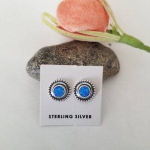 May include: A pair of sterling silver stud earrings with blue opal stones set in a circular bezel. The earrings are displayed on a white card with the words "Sterling Silver" printed on it.