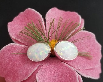 May include: A pair of silver stud earrings with oval-shaped white opal stones. The earrings are displayed on a pink flower.