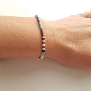 May include: A beaded bracelet with a colorful design. The bracelet features small, cylindrical beads in turquoise, red, black, white, and brown. The bracelet is worn on a wrist.