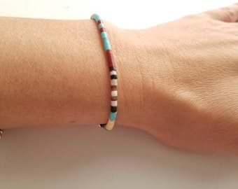 May include: A colourful beaded bracelet. The bracelet is made up of small, cylindrical beads in turquoise, red, black, white, and brown. The bracelet is worn on a wrist.