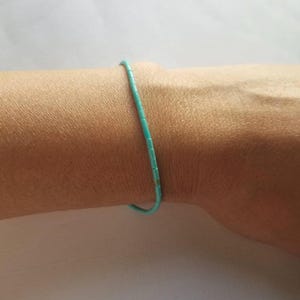 May include: A delicate bracelet composed of small, cylindrical turquoise-colored beads strung together in a single continuous line. The beads are uniform in shape and size, creating a simple, slender band that encircles the wrist with a vibrant color.