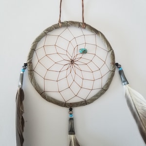 May include: A handmade dreamcatcher with a circular frame wrapped in textured fabric. The web is made of brown string, with a turquoise stone in the center. Feathers and beads hang from the bottom, creating a bohemian aesthetic.
