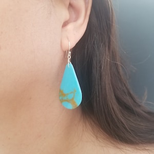 May include: Turquoise dangle earrings with a teardrop shape and gold marbling. The earrings are made with sterling silver.
