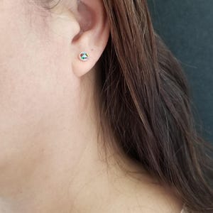 May include: A close-up of a person's ear with a small, colorful, oval-shaped earring. The earring is made of silver and has a variety of colors, including red, green, blue, and yellow.