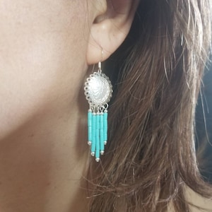 May include: Silver dangle earrings with a turquoise bead fringe. The earrings feature a round silver charm with a floral design.