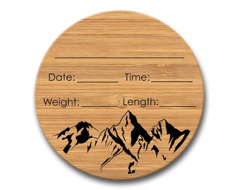 May include: Round wooden plaque with a mountain range design. The plaque has blank lines for "Date", "Time", "Weight", and "Length".
