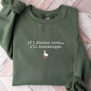 May include: Olive green sweatshirt with the embroidered phrase "if i shenan once... i'll shenanigan" and a small graphic of a goose on a scooter. The sweatshirt is laid flat on a light wood-grain surface.