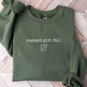 May include: An olive green sweatshirt with the text "engaged girl fall" and a leaf graphic in white. The sweatshirt is folded on a light brown wooden surface.
