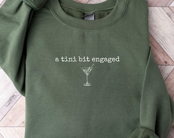 May include: An olive green sweatshirt with the phrase "a tini bit engaged" and a martini glass graphic in white. The long-sleeved crewneck top is made of a soft material and is laid flat on a wooden surface.