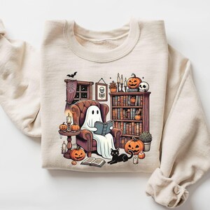 May include: Beige sweatshirt featuring a Halloween-themed illustration. A ghost reads in a cozy armchair surrounded by pumpkins, candles, and a black cat. A bookshelf and a skull add to the spooky ambiance.