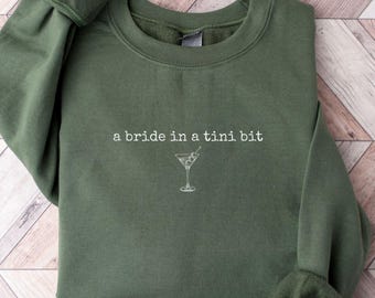 May include: A green sweatshirt with the text "a bride in a tini bit" and a small image of a martini glass.