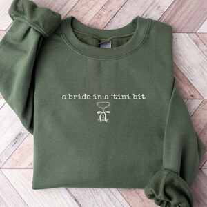 May include: A dark green sweatshirt with the text "a bride in a 'tini bit" and a small illustration of a martini glass.
