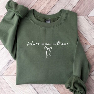 May include: An olive green sweatshirt with the text "future mrs. williams" in a cursive font, along with a bow graphic. The sleeves are cuffed. The sweatshirt is laid on a light brown and white wooden surface.
