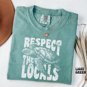 May include: Light green t-shirt with the text "Respect the Locals" and a turtle graphic. The shirt has a crew neck and short sleeves. A silver necklace with a pendant is draped across the shirt.