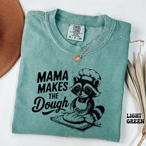 Può includere: Maglietta verde chiaro con una grafica nera di un procione che indossa un cappello da chef e fa l'impasto. Il testo sulla maglietta recita "Mama Makes the Dough".