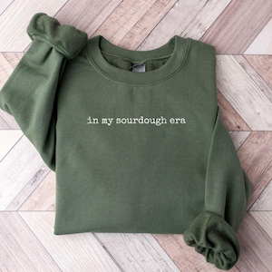 Puede incluir: Una sudadera verde con el texto "in my sourdough era" impreso en blanco en la parte delantera.